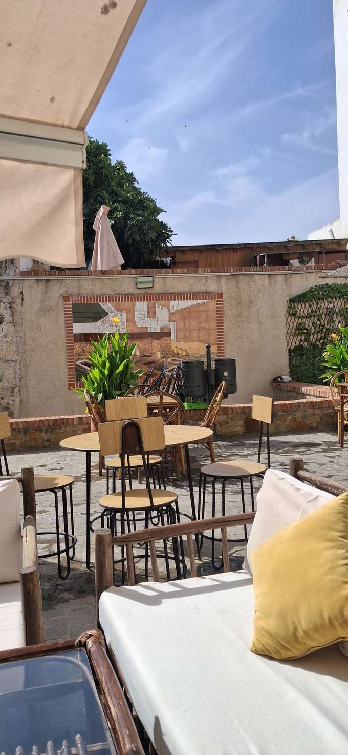 La terrasse de jour : tables et tabourets en bois, banquette à coussins clairs, mur de brique et parasol