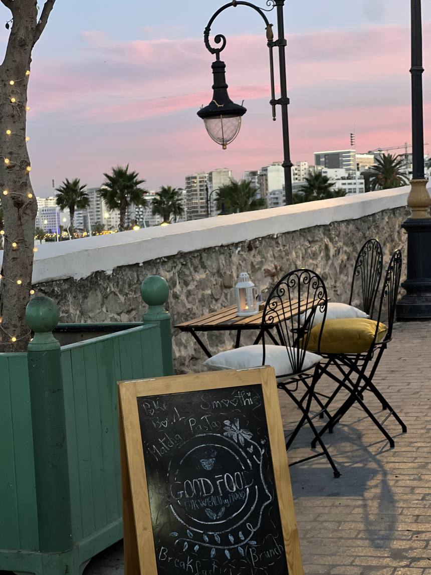 La terrasse au coucher du soleil : chaises en fer forgé et coussins moutarde, guirlandes, ardoise « Good Food », et la ville de Tanger avec ses palmiers en arrière-plan